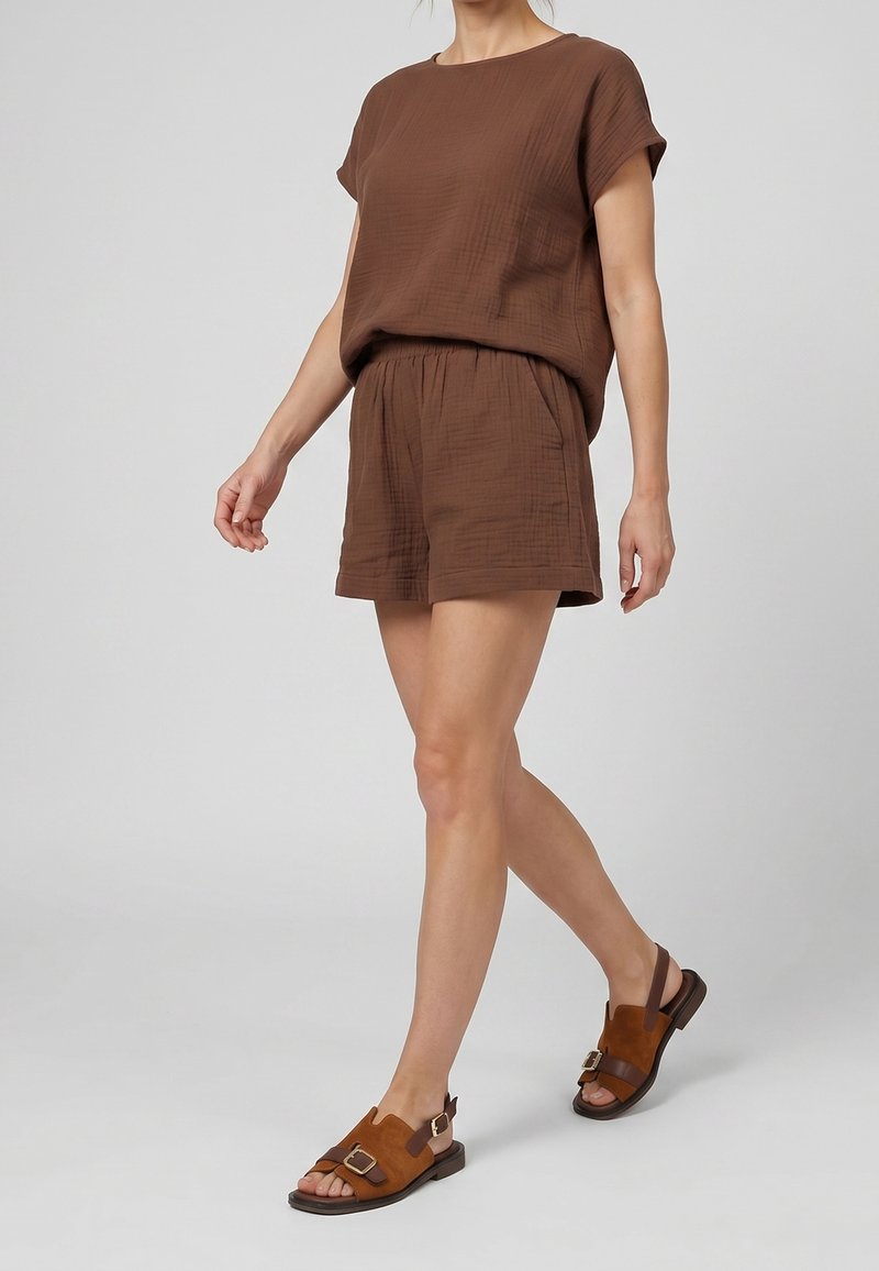 Femme portant un haut marron ample à manches courtes et un short assorti, marchant en sandales marron à bouts ouverts avec des boucles sur un fond gris.