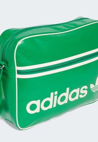 Grønn syntetisk veske med en hvit glidelås og akzentlinjer, med "adidas"-logoen tydelig plassert i hvitt på forsiden.