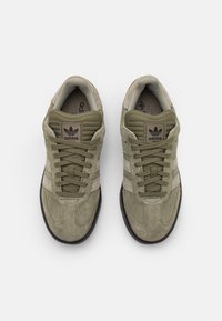 Ett par olivgröna mockaskor från Adidas med beige ränder och svarta sulor, sedda uppifrån mot en vit bakgrund.