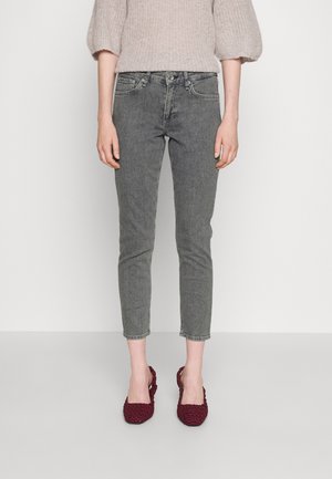 Slim fit vybledlé šedé džíny nošené osobou stojící na šedé podlaze, doplněné béžovým pleteným svetrem a vínově červenými háčkovanými botami.