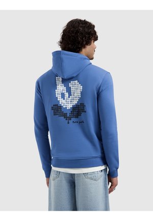 Man draagt een blauwe hoodie met een gepixelde witte en zwarte afbeelding en de tekst "Pure Path" op de achterkant, gecombineerd met lichtblauwe spijkerbroek.