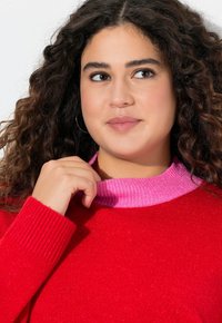 Rode trui met een geribd textuur, met een roze geribde kraag. Model heeft krullerig haar en raakt de kraag met de hand aan.
