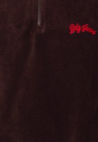 Mørkebrun fleecepullover med kort lynlås og rød broderet "Bdg Jeans"-logo på venstre bryst.