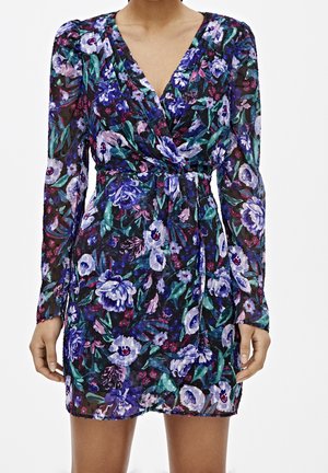 Frau trägt ein kurzärmliges Kleid mit blauem, lila und grünem Blumenmuster und V-Ausschnitt mit geraffter Taille.