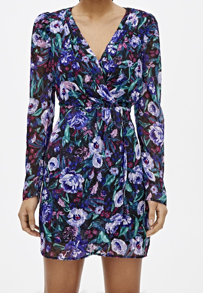 Femme portant une robe courte à manches longues avec un imprimé floral bleu, violet et vert et un décolleté en V avec une taille froncée.