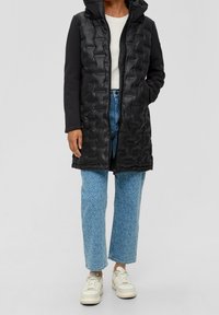 Czarna pikowana kamizelka z kapturem, z wzorem kanapkowym i bocznymi kieszeniami, zestawiona z wzorzystymi jasnoniebieskimi dżinsami i białymi trampkami.