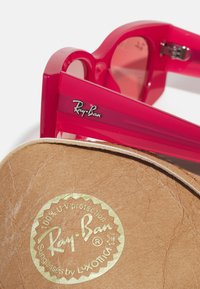 Rosa Ray-Ban solbriller med gjennomsiktige rektangulære rammer og tonede linser, plassert på et brunt etui med et gull UV-beskyttelsestempel.
