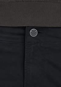 Gros plan de la taille d'un pantalon foncé avec un bouton en métal marqué "PME Legend Jeans" sous l'ourlet côtelé d'un pull gris foncé.
