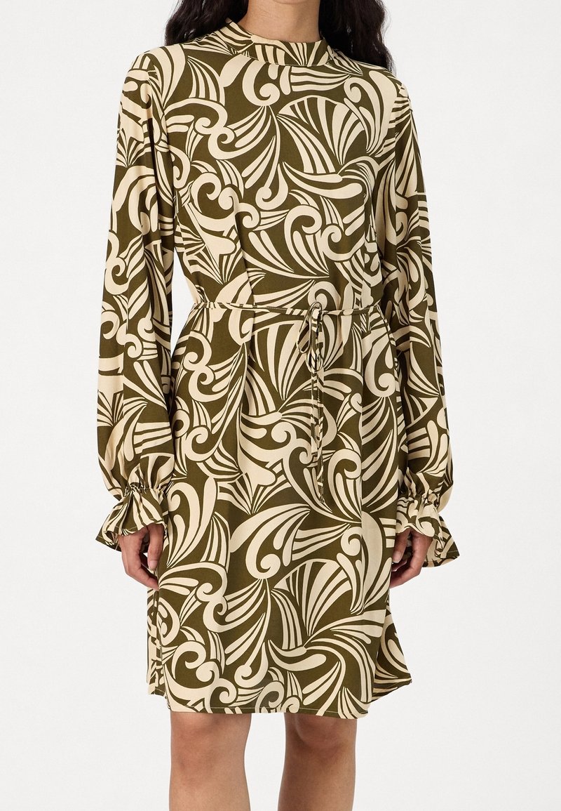 Femme portant une robe mi-longue à manches longues avec des motifs abstraits tourbillonnants olive et beige, ceinturée à la taille.