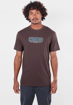 Homme aux cheveux bouclés portant un t-shirt marron avec le logo bleu OXBOW et un pantalon foncé, debout devant un fond clair uni.