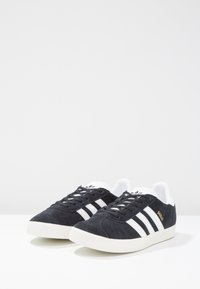 Baskets en suede noir avec trois bandes blanches, bout rond et semelle en caoutchouc blanche. Texte doré "Gazelle" sur le côté. Design classique à lacets.