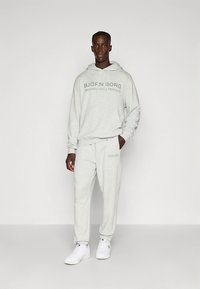 Light gray hoodie and sweatpants set, featuring a front logo print. Material has a smooth texture. Worn with white sneakers.