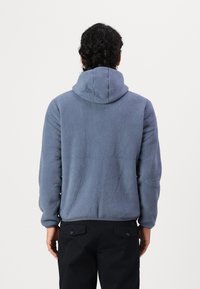 Blue fleece hoodie with a textured exterior, a spacious hood, and ribbed cuffs, worn with black pants featuring two rear pockets.