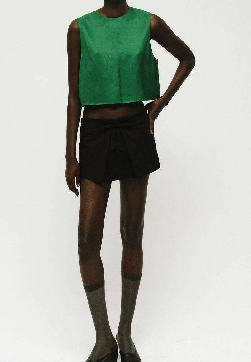 Personne portant un haut court vert sans manches, une mini-jupe noire avec un détail à l'avant, des chaussettes grises montantes jusqu'aux genoux, et des chaussures noires, debout avec une main sur la hanche.