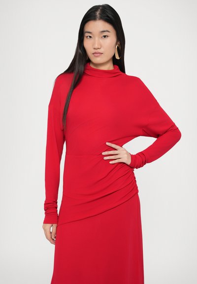 Κόκκινο φόρεμα μακρυμάνικο με λαιμόκοψη τύπου ντραπέ, με ραφές στις πλευρές που δημιουργούν πτυχώσεις και απαλή υφή υφάσματος. Το μοντέλο ποζάρει με το χέρι στη μέση.