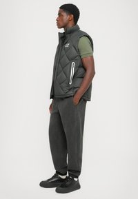 Black puffer vest with quilted design, featuring zippered side pockets and a high collar. Worn over an olive green shirt and gray patterned pants.