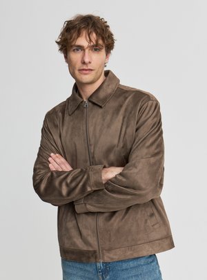 Młody mężczyzna o kręconych włosach, ubrany w brązową zamszową kurtkę zapinaną na zamek z kołnierzem, ze skrzyżowanymi ramionami, stojący na tle jednolitego jasnoszarego tła.