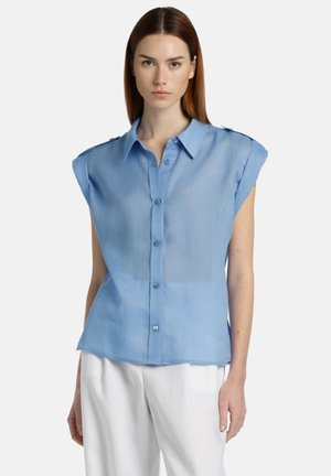 Lichtblauw kortwaardig shirt met knopen aan de voorkant, zachte textuur en schouderaccenten. Gecombineerd met witte broek voor contrast.