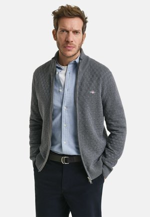 Grijze ritscardigan met een gestructureerd ruitpatroon, geribbelde kraag, gedragen over een lichtblauw shirt en een donkere broek. Met een riem om de taille.