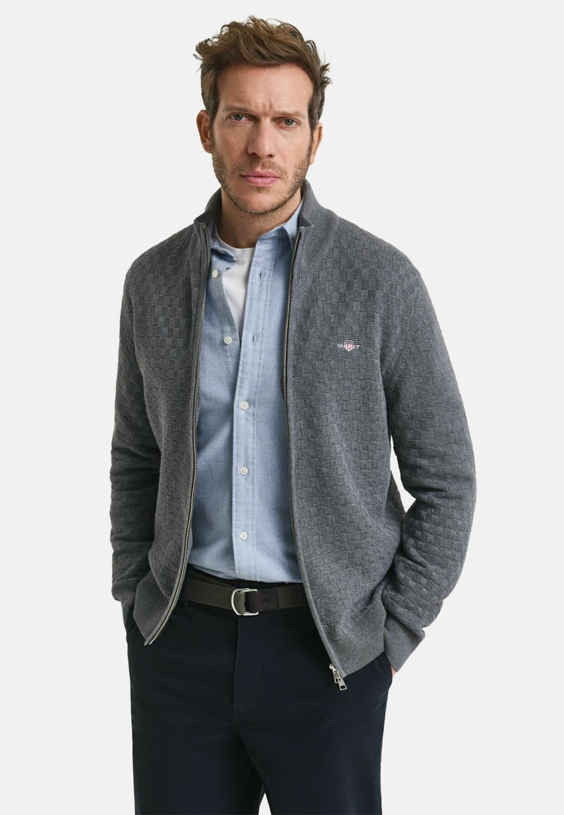 Grauer Reißverschluss-Kardigan mit strukturiellem Karomuster, geripptem Kragen, getragen über einem hellblauen Hemd und dunklen Hosen. Am Bund mit einem Gürtel geschnallt.