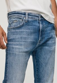 Hombre vestido con jeans descoloridos de un azul claro y una camiseta blanca, con la mano izquierda relajada a su lado contra un fondo sencillo.