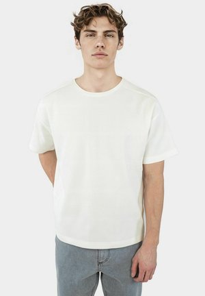 White ribbed t-shirt with a round neck, short sleeves, and a relaxed fit, paired with gray denim jeans. Soft texture and casual design.