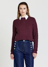Maglione borgogna a maglia con spalle scese, indossato sopra una camicia bianca con colletto. Abbinato a un paio di jeans in denim blu a vita alta con dettagli in bottone.