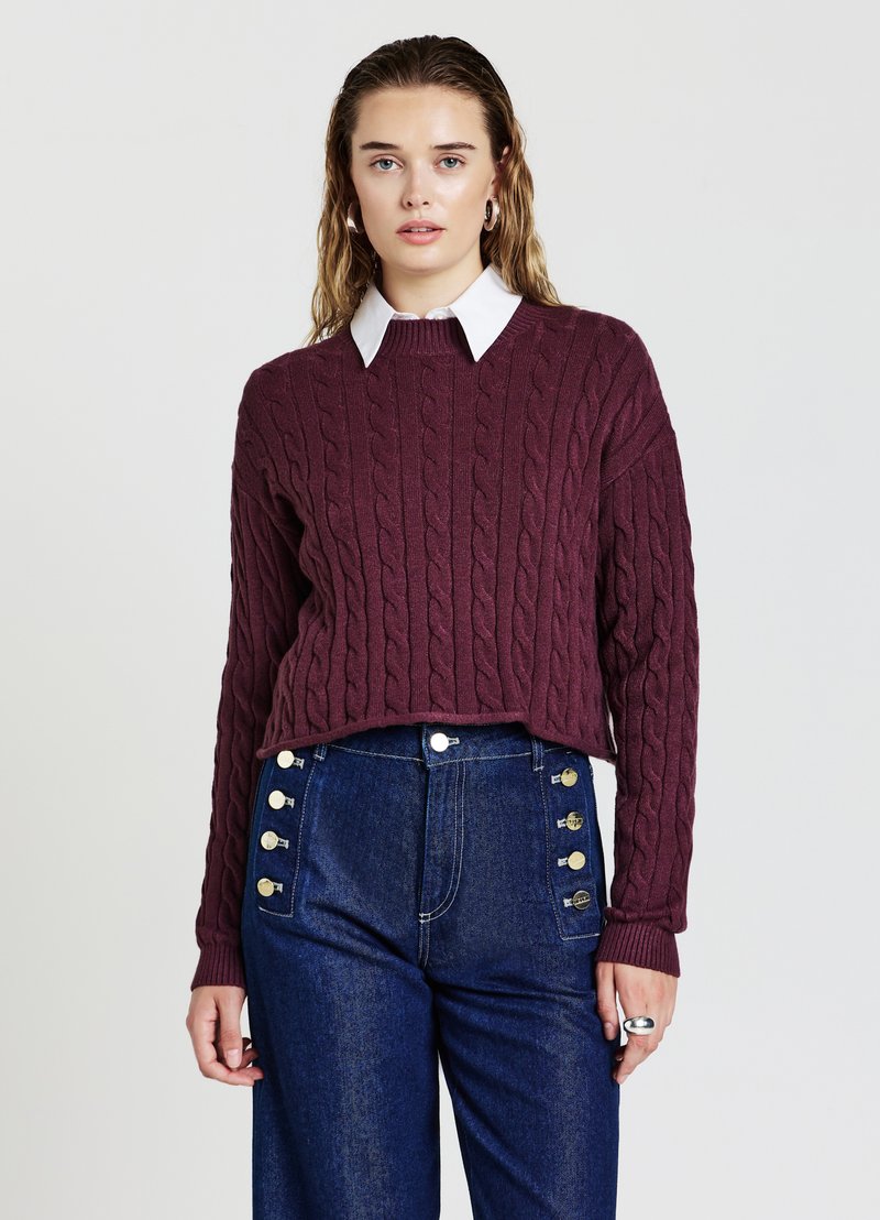 Maglione borgogna a maglia con spalle scese, indossato sopra una camicia bianca con colletto. Abbinato a un paio di jeans in denim blu a vita alta con dettagli in bottone.