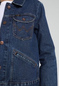 Mörkblå denimjacka med klassisk krage, texturerad tyg, knäppning med knappar, bröstficka med en zigzag-stygn och sidficka.