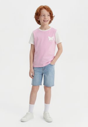 Chlapec s červenými vlasmi v bielych teniskách, bielych ponožkách, rifľových šortkách a ružovobielom tričku s bielym motýľom a japonským textom.