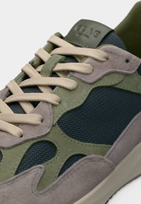 Κοντινό πλάνο πράσινου, γκρι και μπεζ αθλητικού παπουτσιού με κορδόνια μπεζ και λαστιχένια σόλα, που δείχνει υφασμάτινη υφή και σουέντ επενδύσεις.