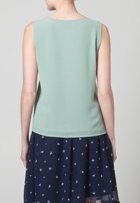 Top senza maniche verde menta con una texture liscia, caratterizzato da un scollo dritto. Abbinato a una gonna blu scuro decorata con motivi di uccelli bianchi.