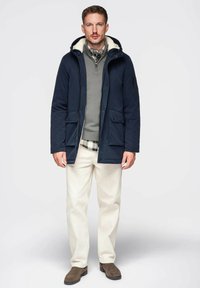 Parka bleu marine avec capuche bordée de fourrure, poches avant, superposée à un pull gris et une chemise à carreaux, associée à un pantalon crème et des bottes marron.