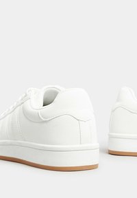 Vita atletiska skor gjorda av syntetiskt material, med en slät yta, rundad bakdel och en ljusbrun gummisula.