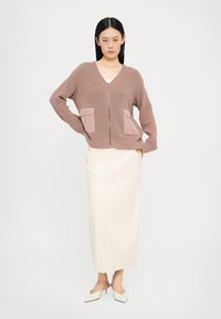 Strikket cardigan i mauve med to frontlommer og glidelås, kombinert med et kremfarget midi-skjørt og hvite spisse sko.