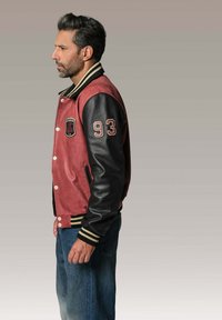 Veste bomber rouge et noire avec des manches en cuir, des poignets côtelés et des patchs brodés incluant "93" sur le bras. Coupe standard, style décontracté.