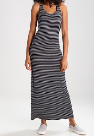 Kobieta w czarno-białej, poziomo paskowanej, bezrękawnikowej sukience maxi, w białych wsuwanych butach, stojąca na jednolitym tle.