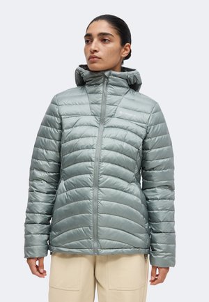 Kvinde iført lysegrå quiltet hættejakke med frontlynlås og beige bukser, stående mod en ensfarvet lys baggrund.