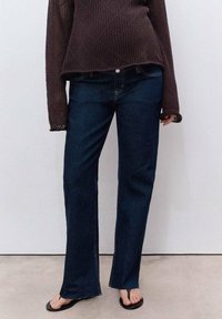 Persona vestida con jeans rectos de color azul oscuro, sandalias negras de punta abierta y un suéter de manga larga de color marrón oscuro frente a una pared lisa.