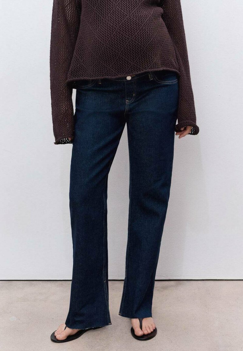 Persona vestida con jeans rectos de color azul oscuro, sandalias negras de punta abierta y un suéter de manga larga de color marrón oscuro frente a una pared lisa.
