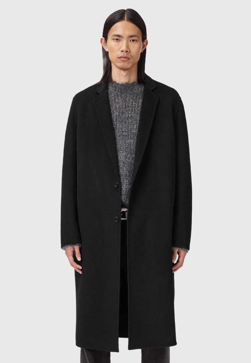 Palton negru din lână cu o croială ajustată, având un guler cu îndoitură, două butoane și buzunare laterale, purtat peste un pulover texturat gri.