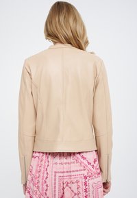 Chaqueta de cuero beige con un diseño ajustado, que presenta acentos acanalados en los hombros y una longitud corta. Puños con cremallera para más detalle.