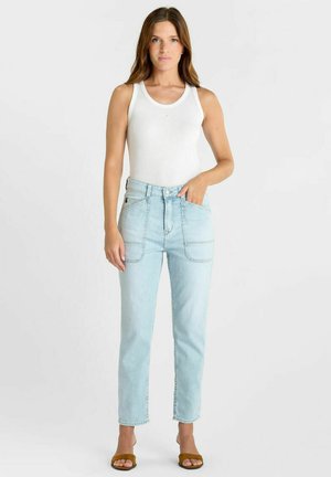 Vrouw met een wit mouwloos topje, lichtblauwe high-waisted jeans met voorzakken, en bruine sandalen met open tenen, staand met één hand in de zak.
