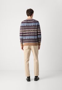 Strickpullover mit geometrischen Mustern in Blau, Braun und Creme, mit gerippten Bündchen und Saum. Beigefarbene Hose und schwarze Schuhe ergänzen das Outfit.