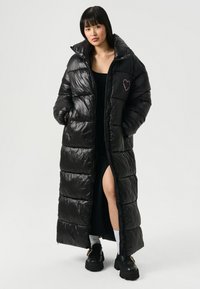 Mujer con un abrigo de plumas largo y brillante negro con un diseño de corazón rosa, vestido negro con una abertura en el muslo, calcetines blancos y zapatos de plataforma negros.
