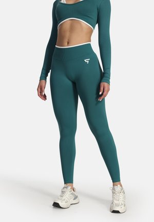 Osoba nosiaca tyrkysové legíny s vysokým pásom a zodpovedajúci crop top s dlhým rukávom a bielymi teniskami, stojaca na bielom pozadí.