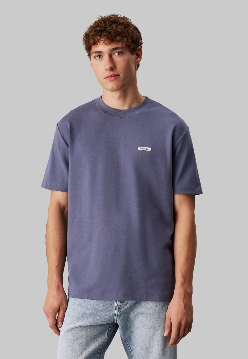 Camiseta de cuello redondo morada de algodón con mangas cortas, que presenta un pequeño parche con el logotipo de Calvin Klein en la parte izquierda del pecho. Ajuste clásico.