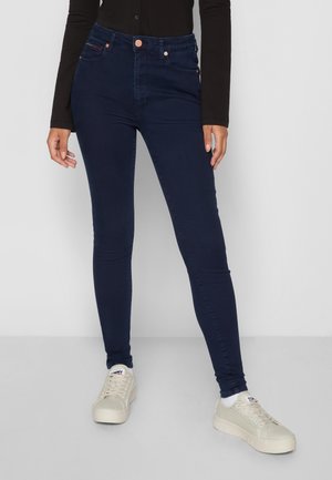 Vrouw draagt donkere blauwe strakke spijkerbroek, witte sneakers en een zwart shirt met lange mouwen, staand op een grijze vloer tegen een witte achtergrond.