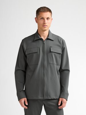 Junger Mann trägt eine dunkelgraue Jacke mit Reißverschluss und zwei Brusttaschen sowie passende Hose und steht vor einem einfachen hellen Hintergrund.