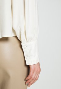 Weißes Oberteil mit langen, gerafften Ärmeln, mit Rüschensäumen und zwei Druckknopfverschlüssen. Glatte Textur; getragen mit beigen Pants.